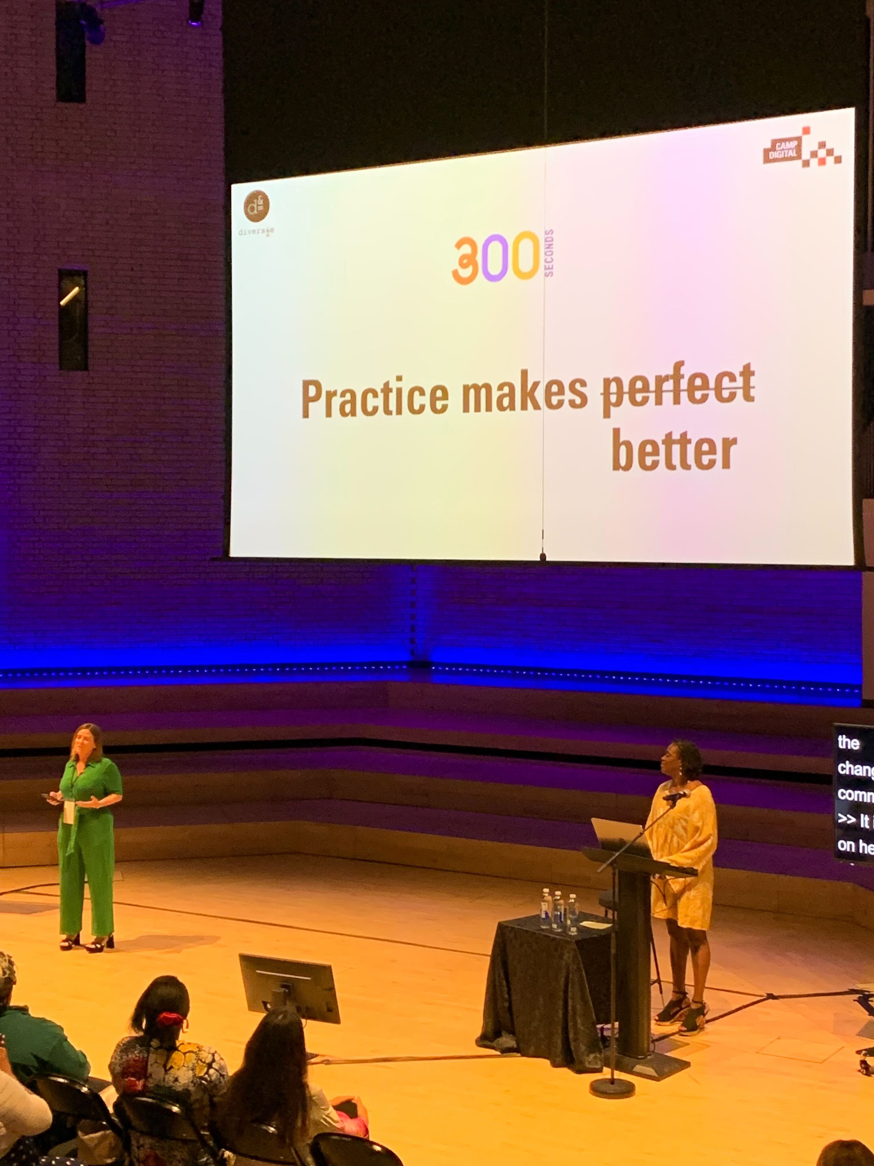 Sharon O'Dea and Annette Joseph stand in front of the audience. The screen behind them reads practice makes perfect. The perfect is crossed out and replaced with better, so it reads practice makes better.